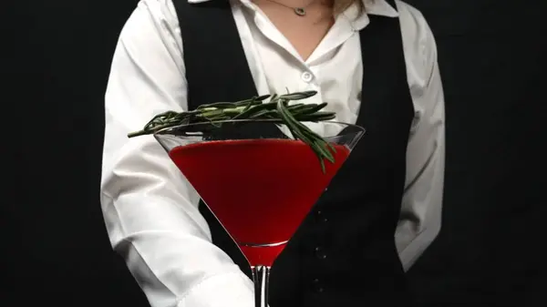 stock image Macrography, witness the artistry of a bartender serving a vibrant red cocktail in a martini glass, beautifully adorned with a sprig of fresh rosemary against a sleek black background. Comestible.