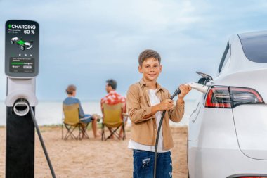 Aile tatili gezisi plajda elektrikli arabayla geziyor, küçük çocuk EV 'yi şarj ederken ailesi deniz manzarasının tadını çıkarıyor. Alternatif enerjili ve çevre dostu arabası olan bir aile gezisi. Kalıcı