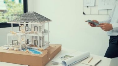 Profesyonel yakışıklı Asyalı proje yöneticisi ev modelini kontrol etmek için tablet kullanırken ev modelinin yapısını dikkatlice incelerken arkasında da planlar var. Yaratıcı tasarım. Tertemiz..