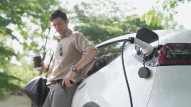 Yavaş çekim adamı ve EV elektrikli araba ile sürdürülebilir şehir ulaşımı ilkbahar bahçesindeki açık kahve kafeteryasında şarj oluyor, yeşil şehir sürdürülebilirliği ve çevre dostu EV aracı. Yararlı