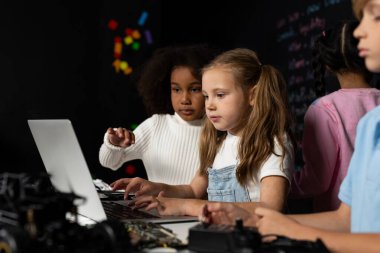 Beyaz önlüklü akıllı kız BTMM sınıfında dizüstü bilgisayar kullanarak robotik teknolojiyi öğreniyor. Mavi tişörtlü öğrenci anakart öğretirken zeki öğrenciler kod okumaya çalışıyor. Etkinlik.