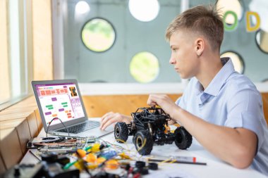 Liseli çocuk ekranda kod izliyor ve STEM sınıfında teknoloji öğrenirken robotik araba tutuyor. Masaya dizüstü bilgisayar, kumanda, elektrik kablosu, şarj aleti ve robot araç koyun. Öğretim.