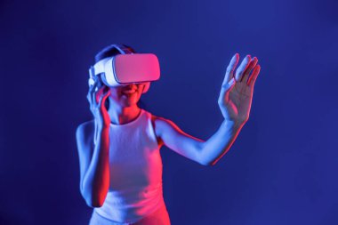 Çevresi siberpunk neon ışıklarıyla çevrili zeki bir kadın metaevrene bağlı VR kulaklık takıyor, gelecekçi siber uzay topluluğu teknolojisi. Sanal objeye el ile dokunan kadın. Halüsinasyon.