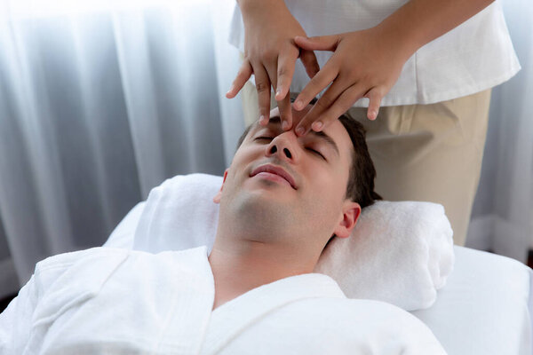 Кавказский мужчина наслаждается расслабляющим анти-стрессовым массажем головы и расслабляющим отдыхом кожи лица в дневном спа современном светлом окружении на роскошном курорте или в спа-салоне отеля. Quiescent