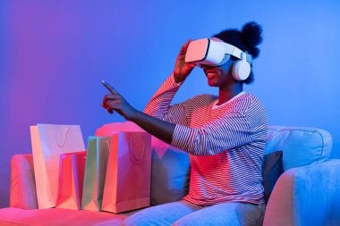 VR 'lı genç Afrikalıların alışverişinin tadını çıkarın. İnternette hologram uygulaması, güzellik ürünleri, neon ışıklandırma, oturma odasındaki koltukta alışveriş torbası bulunan canlı dijital fütüristik teknoloji. Entrika.