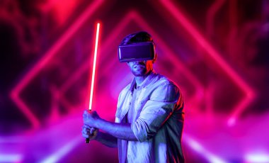 Lazer kılıcı tutan ve VR gözlük takan oyuncu. Beyaz adam, neon şatonun etrafında dikilirken görsel gerçeklik kulaklığı kullanıyor. Yenilik teknolojisi. Sapma.