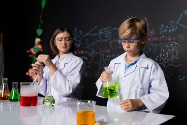 Labaratuvarda yardımcı öğretmen öğrenci laboratuvar önlüğü ve gözlük takıyor ve STEM sınıfında kimya bilimi üzerine deney yapıyorlar. Öğrenci derneği, beher içinde sarı sıvıya indi. Etkinlik.