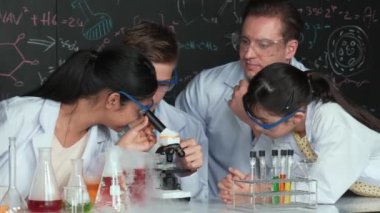 Çok kültürlü bir öğrenci mikroskop altında incelerken güzel bir kız da numuneyi inceliyor. BTMM sınıfında karatahta bilim teorisi deneyi yapan liseli çocuklara bakan öğretmen. Öğretim