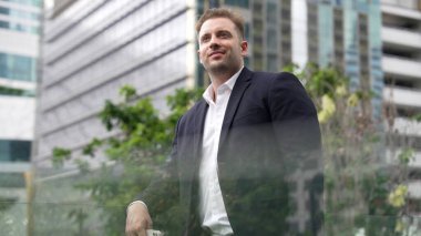 Kafkasyalı iş adamı cep telefonunu tutuyor ve gökdelen manzarasına bakıyor. Modern binanın çatısında duran yetenekli erkek lider. Kafkasyalı yönetici gözlerini dinlendirirken telefona bakar. Urbane.