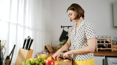 Enerjik anne, mutfak malzemeleri ile çevrili sebzeleri doğrarken modern mutfakta kahvaltı için salata yapıyor. Akıllı kadın temizlikçi yemek pişirir ve yemek hazırlar. Sağlıklı bir yaşam tarzı. Pedagoji.
