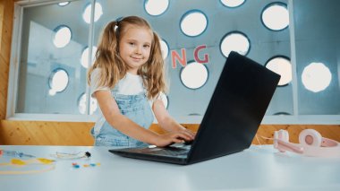 Çalışmak ya da oyun oynamak için dizüstü bilgisayar kullanan küçük tatlı bir kız. Öğrenci kameraya bakıp gülümserken bilgisayarda çalışıyor. Beyaz çocuk kulaklık kullanarak çevrimiçi derse katılıyor. Etkinlik.