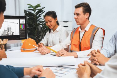 Mimari proje üzerinde birlikte çalışan çeşitli inşaat mühendisi ve müvekkil grupları, inşaat planını gözden geçirip toplantı masasında plan oluşturuyorlar. Tedbirli