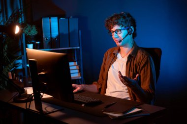 Genç yaratıcı iş adamı, bilgisayar ekranında dinleyicilerle iletişim kuruyor ya da takım üyesiyle görüşüyor. Neon Light Modern Ev Ofisi 'nde kulaklık takıyor. Gece vakti çalışma kavramı. Toplayıcı.