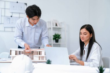 Profesyonel Asyalı erkek mimar ev modelinin uzunluğunu ölçmek için cetvel kullanırken, beyaz tenli genç meslektaşı ev modeliyle toplantı masasındaki verileri analiz etmek için dizüstü bilgisayar kullanıyor. Tertemiz..