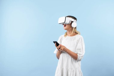 Kafkasyalı bir kız elinde telefon tutarken ve görüş açısını gösterirken VR gözlük takıyor. Görsel gerçeklik dünyasını ya da metaevreni keşfetmek isteyen mutlu kadın şaşırdı. Teknoloji yeniliği. Kısıtlanma.