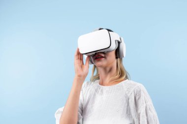 Mutlu kadın VR gözlüklerine bakıp mavi arka planda dururken şaşırdı ya da heyecanlandı. Beyaz pijamalı beyaz bir kız metaevrene bağlanmak için görsel gerçeklik gözlüğü kullanıyor. Kısıtlanma.