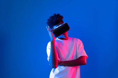 Sanal gerçekliği dijital fütüristik teknolojiye bağlayan pembe mavi neon duvarda VR 'dan bakan genç Afrikalı Amerikalı' ya şaşırdım. Entrika.