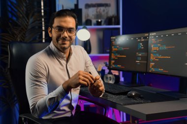 Bilgisayar ekranındaki yazılım geliştirme kodlamasına karşı poz vermek için akıllı bilişim geliştirici görünümlü kamera profilleri, neon ışıklı siber oda ofisinde program güncellemesi internet sitesi verileri sunuyor. Tahmin et..