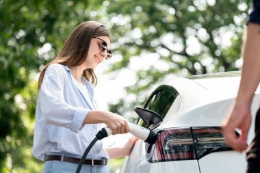 Doğal orman ya da milli parktaki araba yolculuğu sırasında elektrikli araba için şarj olan güneş gözlüğü takan sevimli genç bir çift. Tatil ve tatil boyunca çevre dostu seyahat. Yükselt