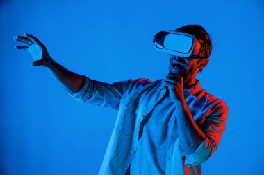 Sanal programı keşfetmek ve girmek için VR gözlük takan heyecanlı bir adam. Yetenekli oyuncular metaevrene dokunurken gündelik giysiler giyer ve neon ışıklı arka planda dururlar. Yaşam tarzı. Sapma.