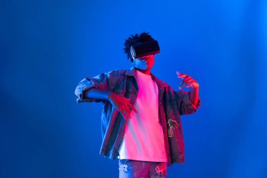 Gülümseyen Afrikalı genç Amerikalı sanal gerçekliği dijital fütüristik teknolojiye bağlayan pembe mavi neon duvarda hologram oyunlarıyla VR dansına bakıyor. Entrika.