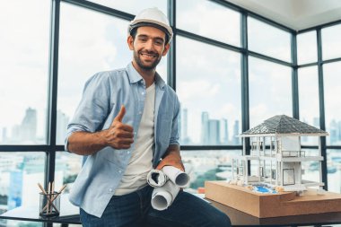 Modern ev projesini düşünen profesyonel mimar mühendisinin portresi güvenlik kaskı ve günlük kıyafetler giyerken. Akıllı iç mimar ev modelinin yanında proje planları tutuyor. İzleme.