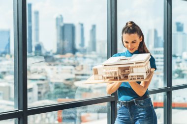 Güzel genç mimar mühendisi ev modelini incelerken mimari modeli elinde tutuyor. Şehir manzaralı pencerenin yanında dururken profesyonel iç mimar ev inşaatını kontrol ediyor. İzleme.