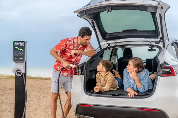 Aile gezisi plajda elektrikli arabayla seyahat ediyor, güzel bir aile bagajda oturuyor, EV araba aküsünü yeşil ve sürdürülebilir enerjiyle şarj ediyor. Aile seyahati ve çevre dostu araba. Kalıcı