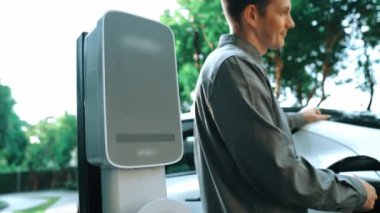 Yavaş çekim adamı yeşil sürdürülebilir şehir parkı yaz bahçesinde EV elektrikli otomobilini kentsel sürdürülebilirlikteki gençlerin yaşam tarzı olarak şarj et.