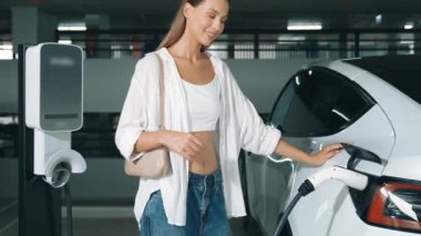 Yavaş hareket eden kadın mutlu ve EV elektrikli arabasıyla şehir merkezindeki Shopp Center otoparkına doğru gülümsüyor. Elektrikli taşıt iç organları için EV şarj prizi taşıyor.