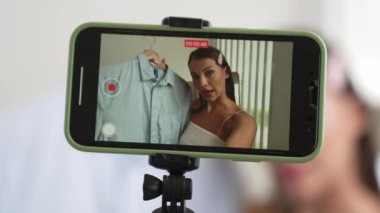 Genç sosyal medyum kadın moda videosu yapıyor. Blogcu kameraya selfie çubuğu ve ışık halkası kullanarak gülümserken, aynı zamanda ikna edici çevrimiçi kıyafetlerin izleyicilere veya takipçilere vlog satmasını sağlıyor. Düzenle