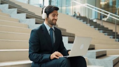 Kendine güveni tam bir iş adamı başarılı iş fikrini müzik dinlerken kutluyor. Happy yönetici projesi kulaklık ve takım elbise giyerken dizüstü bilgisayarla çalışıyor. Yatırımcı merdivenlerde oturuyor. Sevinçli..
