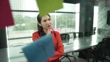 Profesyonel genç ve stresli iş kadını pazarlama sorunlarını çözmenin yollarını düşünürken aynı zamanda renkli yapışkan notlarla ve pazarlama fikriyle cam tahtada duruyor. Yönlendirici.