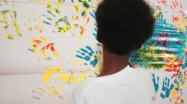 Mutlu Afrikalı çocuğun arkadan bakışı, resim dersinde günlük beyaz gömlek giyerken boyalı duvarı renkli eliyle boyadı. Mart öğrencisi yaratıcı sanat çalışması yapmak için el izi kullanıyor. Eğitim. Öğretim
