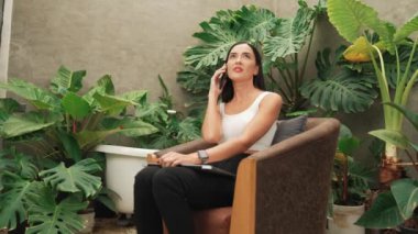 Modern genç kadın telefonda konuşuyor, uzaktan çalışıyor ya da tropik bitkilerle birlikte minimalist bir mimari beton stil yaz egzotik bitki yetiştirme bahçesinde yalnız başına dinleniyor. Blithe