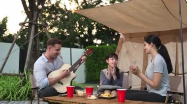 Baba gitar çalarken anne ve kız birlikte kampta oturuyorlar. Ebeveynler, genç nesil çapraz kuşak farklılıklarıyla zaman geçirmek için açık hava kamp aktivitelerini kullanıyorlar. Uyumsuzluk.