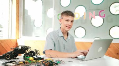 Akıllı genç dizüstü bilgisayarda çalışıyor ve BTMM teknoloji sınıfında kameraya bakıyor. Beyaz tenli bir öğrenci bilgisayar kullanarak verileri analiz ederken masada araba modelinin yanında gülümsüyor. Öğretim