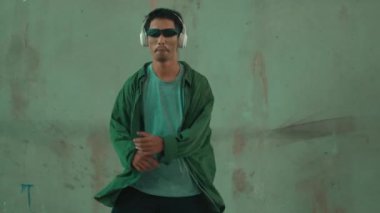 Hippi, şehir caddesinde hip-hop dinlerken kulaklık takıyor. Sokak dansçısı ayak seslerini kulaklıktan müziğe taşıyor. Açık hava sporu 2024. Çimento arka planı. Moda. Aktif stil. hiphop.