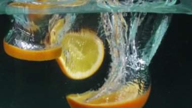 Arkaplanlı cam bir kase suda yüzen portakal dilimleri. Portakal dilimlenmiş makrografi. Ayrı bir karanlık zemin ile berrak suya düşüyor. Kapatın. Ağır çekim. Pabulum.