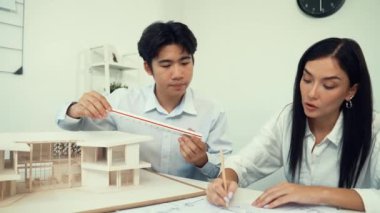 Ev modeli uzunluğunu ölçen profesyonel Asyalı mimar mühendisi, modern ofisteki dizüstü bilgisayar analiz verilerini kullanarak plan üzerine yazan Kafkasyalı iş arkadaşıyla birlikte çalışıyor. Tertemiz..