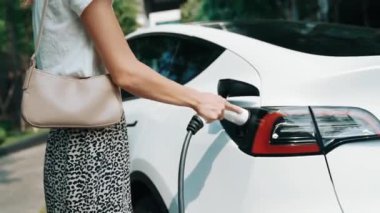 Genç kadın EV elektrikli araba aküsünü şehir merkezindeki EV şarj istasyonunda elektrik iç organları için sürdürülebilir yeşil ve yenilenebilir temiz enerji yaşam tarzıyla şarj ediyor.