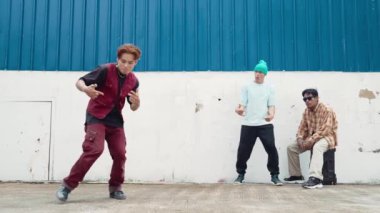 Genç Asyalı dansçı hip hop tarzında dans ediyor ve çok kültürlü arkadaşları duvarın arkasında tezahürat yapıyor. Çekici yakışıklı adam mavi arka planda kollarını uzatıyor. Açık hava sporu 2024 hiphop..