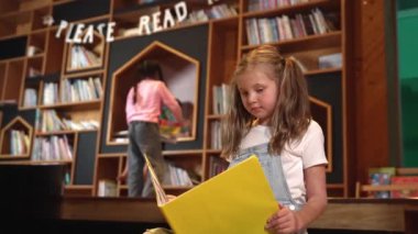 Genç, zeki, beyaz bir kız kütüphanede otururken kitap okuyor. Zeki çocuk öğrenme, okuma, kütüphanede kitap açma. Etkileyici çocuk geçmişi bulanık sayfayı çeviriyor. Etkinlik.