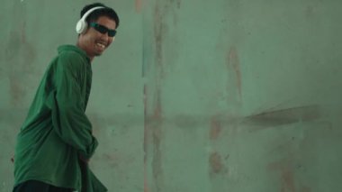 Hippi, şehir caddesinde hip-hop dinlerken kulaklık takıyor. Sokak dansçısı ayak seslerini kulaklıktan müziğe taşıyor. Açık hava sporu 2024. Çimento arka planı. Moda. Aktif stil. hiphop.