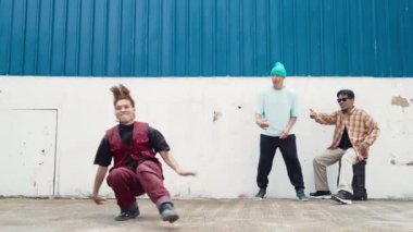 Genç Asyalı dansçı hip hop tarzında dans ediyor ve çok kültürlü arkadaşları duvarın arkasında tezahürat yapıyor. Çekici yakışıklı adam mavi arka planda kollarını uzatıyor. Açık hava sporu 2024 hiphop..