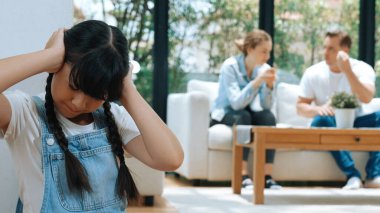 Stresli ve mutsuz genç kız köşeye kıvrılıp kulaklarını kapatarak arka planda ebeveyninin tartışmasını engelliyor. Evde aile içi şiddet ve travmatik çocukluk depresyona dönüşür. Eşzamanlama