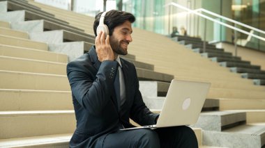 Profesyonel bir iş adamı dizüstü bilgisayarla çalışırken merdivenlerde oturuyor. Yetenekli proje yöneticisi kulaklıktan müzik dinliyor ve e-postaları kontrol ediyor ve pazarlama planı hakkında tartışıyorlar. Açık havada. Sevinçli..