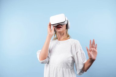 Mutlu kadın VR gözlüklerine bakıp mavi arka planda dururken şaşırdı ya da heyecanlandı. Beyaz pijamalı beyaz bir kız metaevrene bağlanmak için görsel gerçeklik gözlüğü kullanıyor. Kısıtlanma.