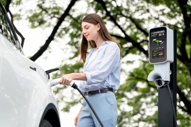 Yolculuk sırasında doğal orman ya da milli parktaki elektrikli araba için şarj olan genç bir kadın. Tatil ve tatil boyunca çevre dostu seyahat. Yükselt