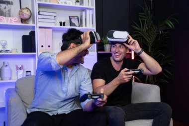 Oyun arkadaşı oyuncular video oyunu oynuyorlar. Oyun kumandası ve sanal teknoloji kulaklığı kullanıyorlar. Stüdyo odasında neon mavi ışıkla. Neşeli dövüşün galibiyle evde rahat bir yaşam. Satılabilir.